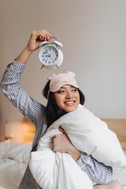 Linda chica sosteniendo un despertador sobre su cabeza y con una sonrisa abrazando una almohada blanca