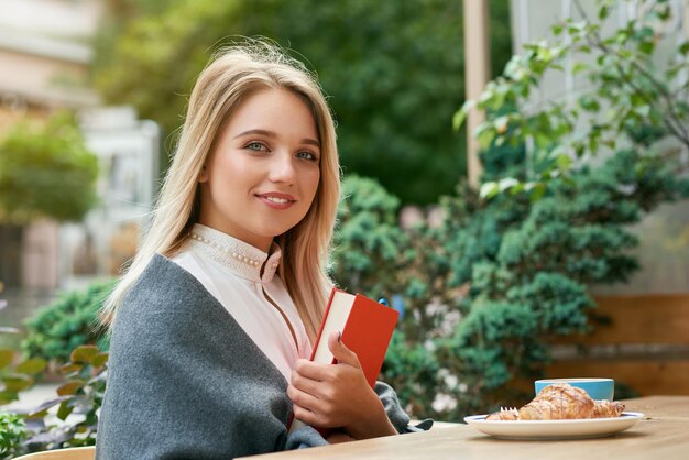 Linda chica sosteniendo gran libro rojo sentado café al aire libre comiendo croissant