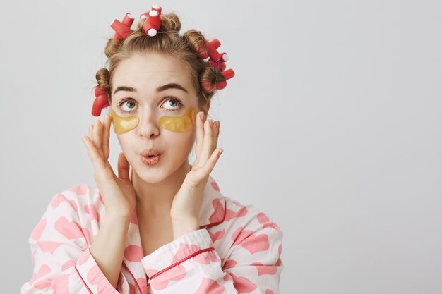 Linda chica tonta en rizadores de pelo y ropa de dormir aplicar parches en los ojos