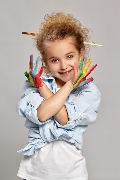 Linda colegiala con un cepillo en su elegante cabello rubio rizado, vestida con una camisa azul y una camiseta blanca. Se cruzó de brazos pintados, sonriendo y mirando a la cámara sobre un fondo gris.