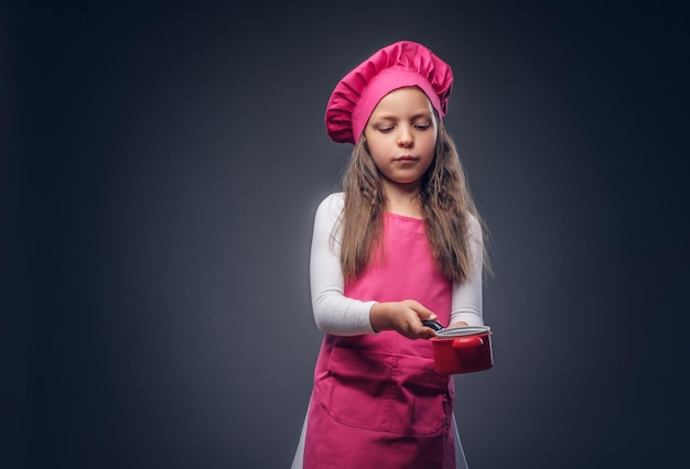 Foto gratuita linda colegiala hermosa vestida con un uniforme de cocinero rosa tiene utensilios de cocina en un estudio. aislado en un fondo oscuro.