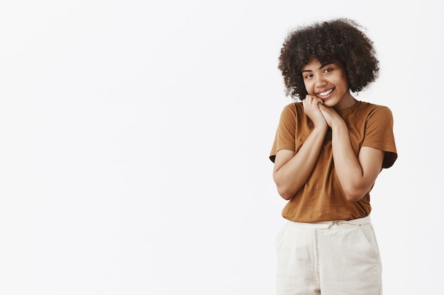 Linda y conmovida mujer afroamericana con cabello rizado en camiseta marrón inclinando la cabeza y apoyándola en las manos sonriendo con expresión encantada y complacida mirando con afecto