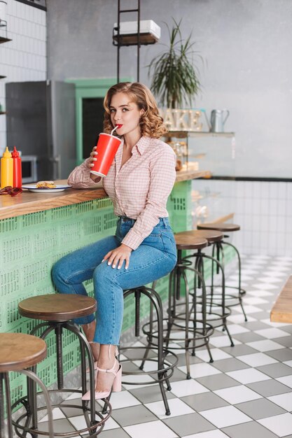 Linda dama con camisa y jeans sentada en el mostrador del bar y bebiendo agua de soda mientras mira felizmente a la cámara en el café