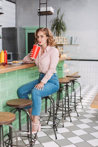 Linda dama con camisa y jeans sentada en el mostrador del bar y bebiendo agua de soda mientras mira felizmente a la cámara en el café