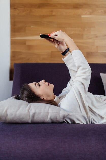 Linda dama en camiseta blanca usando un teléfono inteligente acostado en una cama en casa