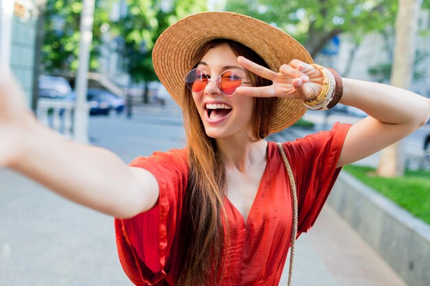 Linda dama con elegante sombrero haciendo selfie mientras camina al aire libre en los fines de semana de verano