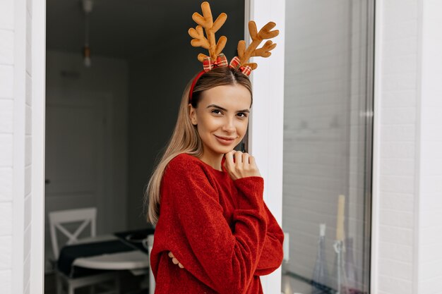 Linda dama encantadora en sombrero de Navidad con suéter rojo posando en casa y preparándose para la celebración de vacaciones