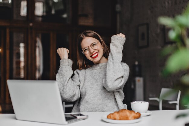 Linda dama con gafas disfruta del éxito, se ríe mientras está sentada en la cafetería con una computadora portátil gris y un apetitoso croissant.