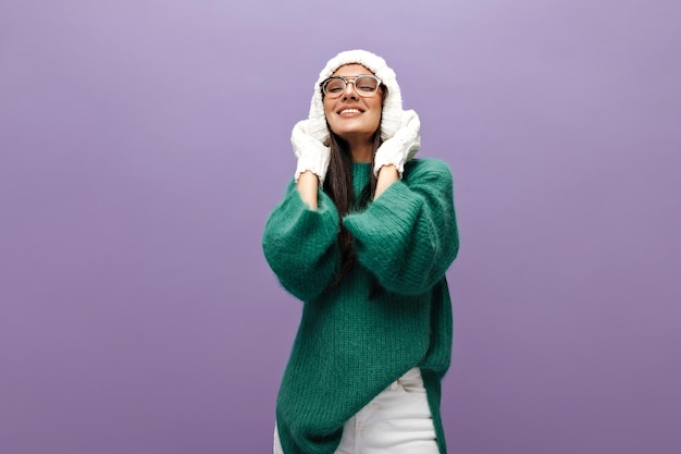 Foto gratuita linda dama con gorro de punto chica morena con guantes blancos, anteojos y suéter verde sonríe sobre fondo púrpura