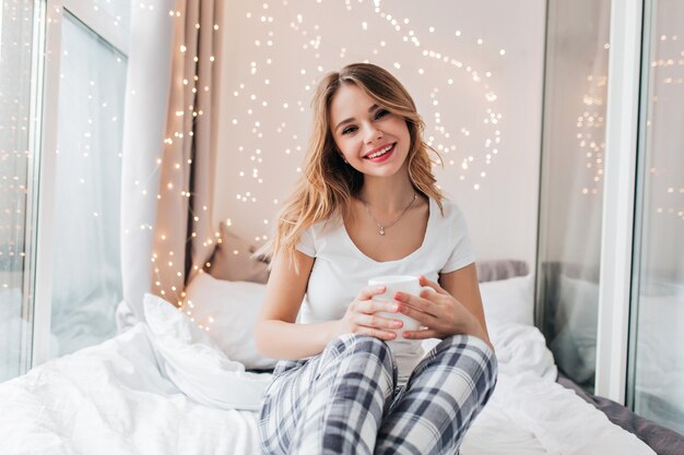 Linda dama rubia en pijama posando en el dormitorio y bebiendo té. Chica increíble con interés y sosteniendo una taza de café.