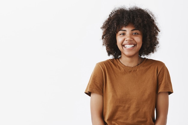 Linda, despreocupada y amigable adolescente afroamericana con peinado afro sonriendo ampliamente con expresión tímida y feliz conociendo nuevos compañeros de clase