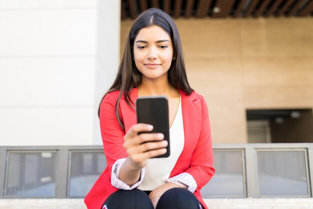 Linda ejecutiva leyendo notificaciones entrantes en el teléfono inteligente mientras está sentada fuera de la oficina