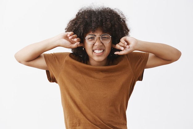 Foto gratuita linda empollona guapa con peinado afro en gafas transparentes prescritas frunciendo el ceño y arrugando la nariz apretando los dientes por el disgusto y la incomodidad cubriendo las orejas sin escuchar ruidos molestos