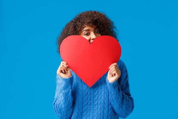 Linda y encantadora niña afroamericana ruborizada, con corte de pelo afro, en suéter, escondiendo la cara detrás de un gran corazón rojo y mirando con alegría, pared azul.