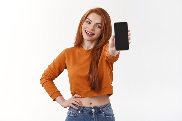 Linda estudiante pelirroja caucásica sonriente en suéter naranja, rastreando su pedido en línea, mostrando una foto genial que quiere publicar en las redes sociales, sosteniendo un teléfono inteligente, pared blanca