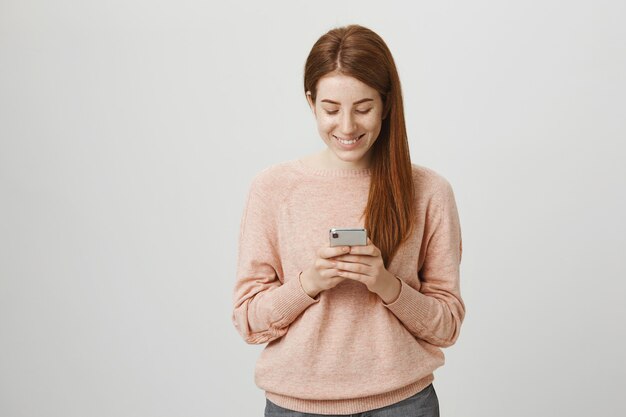 Linda estudiante pelirroja con teléfono móvil y sonriendo a la pantalla