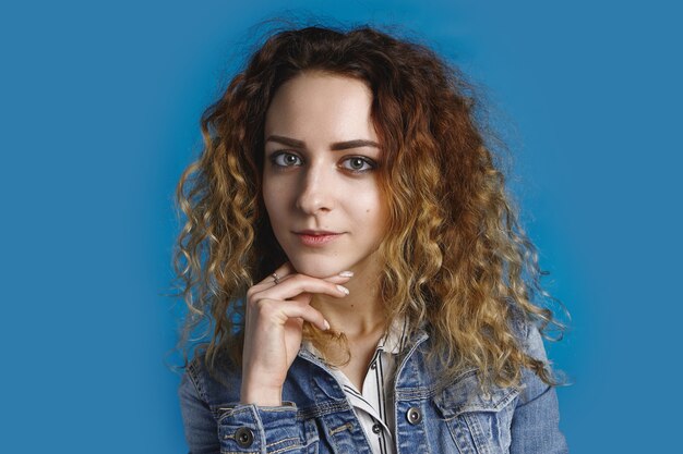 linda estudiante con una piel limpia perfecta, maquillaje natural y peinado rizado, mirando y sonriendo, manteniendo la mano debajo de la barbilla. Concepto de juventud, belleza, estilo y moda