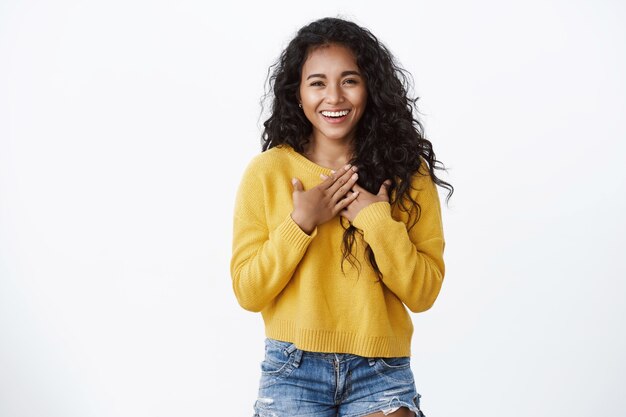 Linda y halagada niña sonriente de cabello rizado, usa un suéter amarillo acogedor, presiona las manos contra el pecho, se siente agradecida y agradece por la ayuda, sonríe aprecia el hermoso regalo