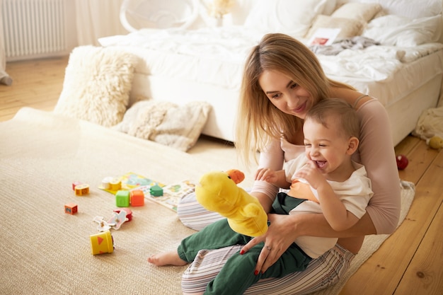 Linda hermana pasar tiempo con su hermanito, sentada en el suelo en el dormitorio. Hermosa niñera joven jugando con el niño en el interior, sosteniendo un pato de peluche. Infancia, puericultura y maternidad