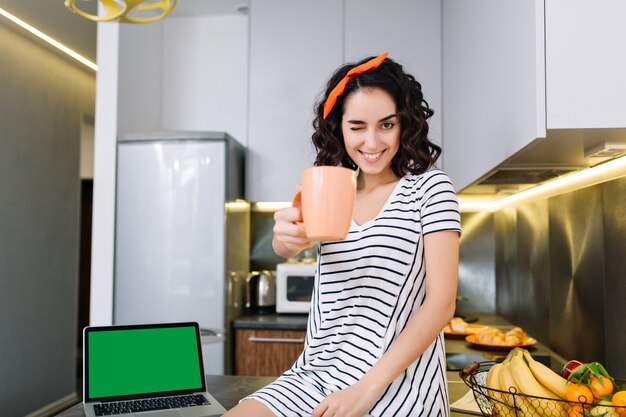 Linda hermosa joven con corte de pelo rizado sonriendo con una taza de té en la cocina en el apartamento moderno. Buenos días, comodidad en casa, fines de semana, divertirse