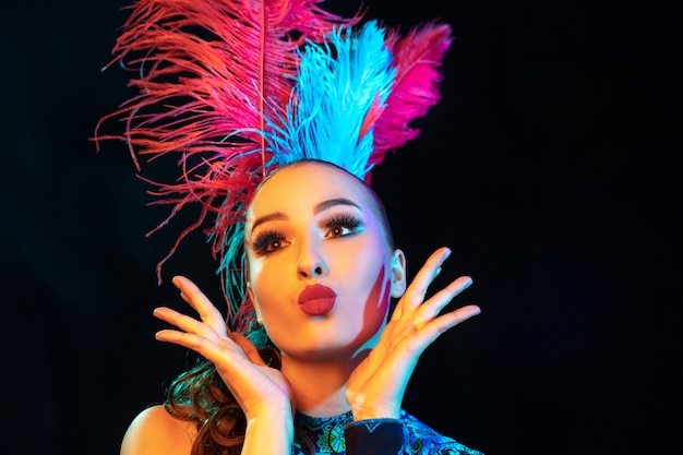 Linda. Hermosa mujer joven en carnaval, elegante disfraz de mascarada con plumas en pared negra en luz de neón. Copyspace para anuncio. Celebración de fiestas, baile, moda. Tiempo festivo, fiesta.