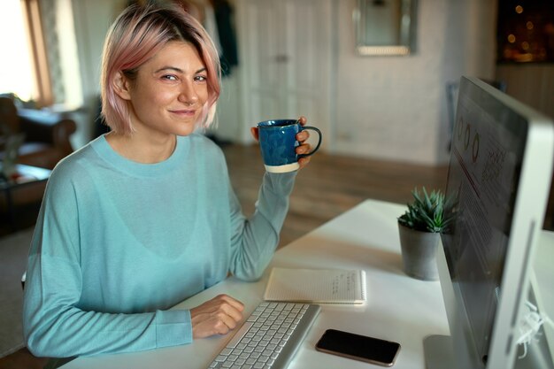 Linda joven diseñadora gráfica trabajando en contenido visual para el sitio web, usando una computadora de escritorio, tomando té, sonriendo a la cámara