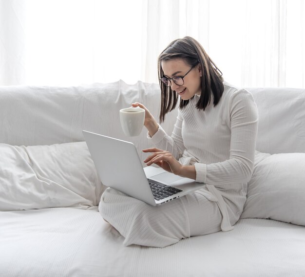 Una linda joven está sentada en su casa en un sofá blanco con un vestido blanco frente a una computadora portátil