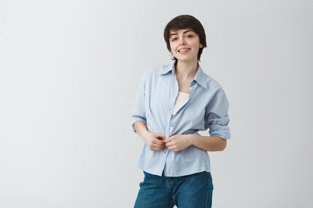 Linda joven estudiante con cabello corto y oscuro sonriendo alegremente, abotonándose la camisa y mirando con expresión feliz y segura.