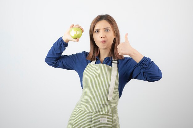 una linda joven modelo en delantal con una manzana verde fresca mostrando un pulgar hacia arriba.