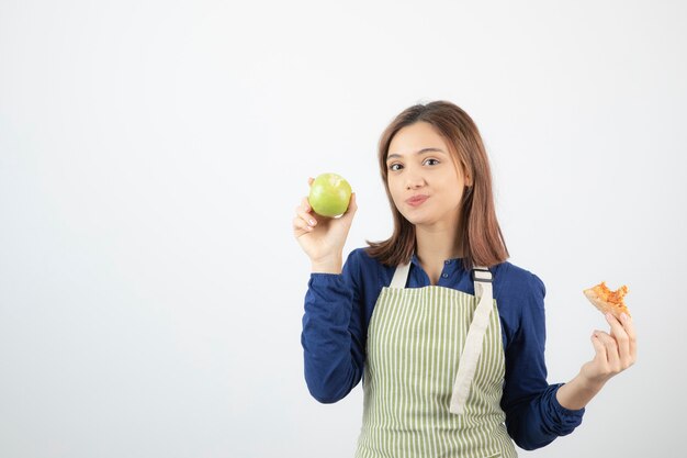 una linda joven modelo en delantal sosteniendo una manzana y un trozo de pizza.