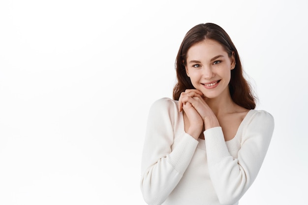 Linda joven modelo femenina se ve sincera y complacida tomándose de la mano cerca de las mejillas y sonriendo a la cámara siendo elogiada mirando agradecida y encantada de pie contra el fondo blanco