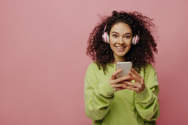 Linda joven morena caucásica con auriculares mira a la cámara sobre fondo rosa Belleza rizada con chaqueta verde sostiene el teléfono en sus manos Concepto de tecnología y personas