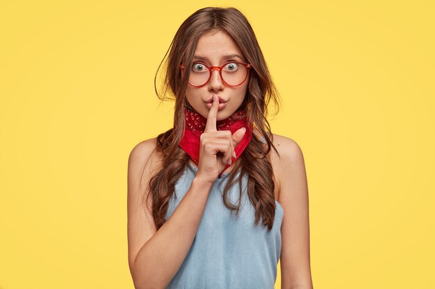 Linda joven morena con gafas posando contra la pared amarilla