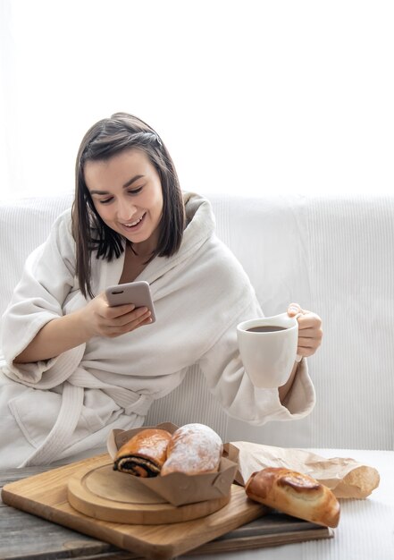 Una linda joven se sienta en el sofá con una bata y toma fotografías de bollos y café. Concepto de brunch y fin de semana.