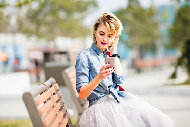 Linda jovencita rubia con pelo corto sentada en un banco de madera y mirando el teléfono inteligente con camisa azul denim y falda de tul gris peinándose el pelo detrás de la oreja.