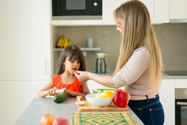 Linda mamá e hija cocinando ensalada para cenar juntos, cortando verduras en la encimera de la cocina, probando una rodaja de pimienta. Tiro medio. Concepto de cocina familiar