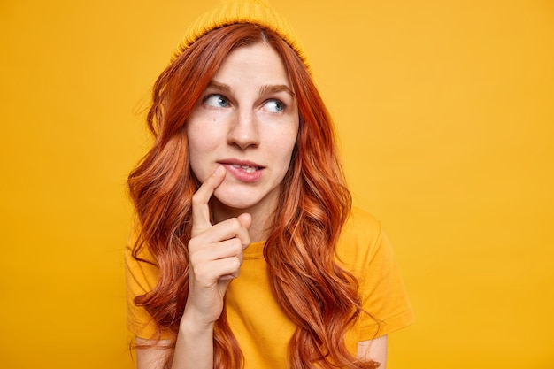 Linda modelo femenina de jengibre de ojos azules mantiene el dedo cerca de los labios intenta tomar una decisión centrada en la derecha contempla algo con sombrero