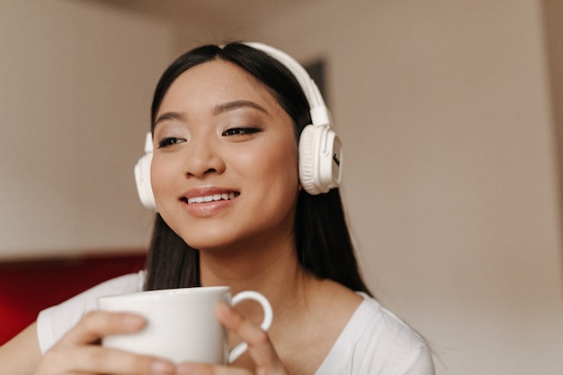 Linda mujer asiática en auriculares grandes está sonriendo y sosteniendo una taza de té