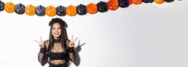 Foto gratuita linda mujer asiática feliz disfrutando de la fiesta de halloween mostrando gesto de paz y sonriendo con costo de bruja