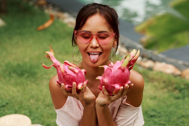 Linda mujer asiática mostrando la lengua, haciendo muecas y sosteniendo las frutas del dragón rosa en las manos.