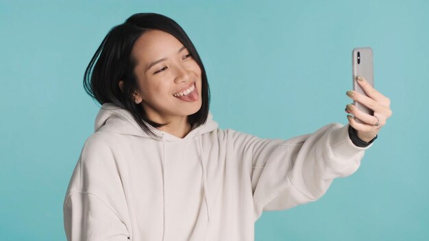 Una linda mujer asiática sacando la lengua haciendo un divertido selfie usando un smartphone aislado de fondo azul