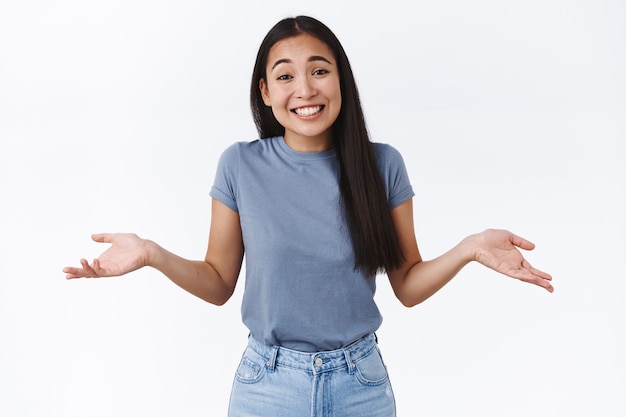 Linda mujer asiática sonriente, despreocupada y relajada, encogiéndose de hombros, inclinando la cabeza tontamente y extendiendo las manos hacia los lados de manera indestructiva, no me importa, no tengo idea y no me voy a preocupar por eso, de pie descuidadamente en la pared blanca