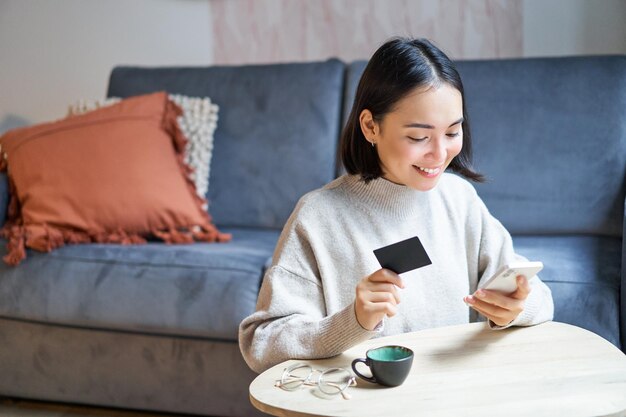 Linda mujer asiática sonriente usando tarjeta de crédito y teléfono inteligente pagando facturas en línea sosteniendo el teléfono móvil l