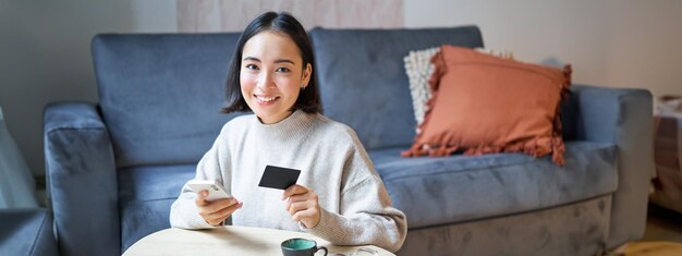 Foto gratuita linda mujer asiática sonriente usando tarjeta de crédito y teléfono inteligente pagando facturas en línea sosteniendo un teléfono móvil