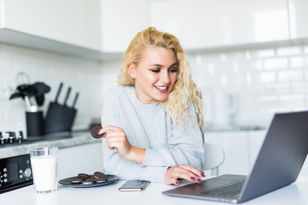 Linda mujer bebiendo leche de un vaso, comiendo galletas usando la computadora portátil mientras está sentado en la mesa de la cocina