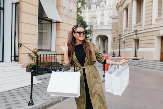 Linda mujer bronceada en elegantes gafas de sol caminando por la calle con paquetes de la tienda