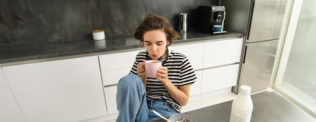 Foto gratuita linda mujer bruneta estudiante desayunando por la mañana bebiendo té caliente sonriendo y pareciendo satisfecha