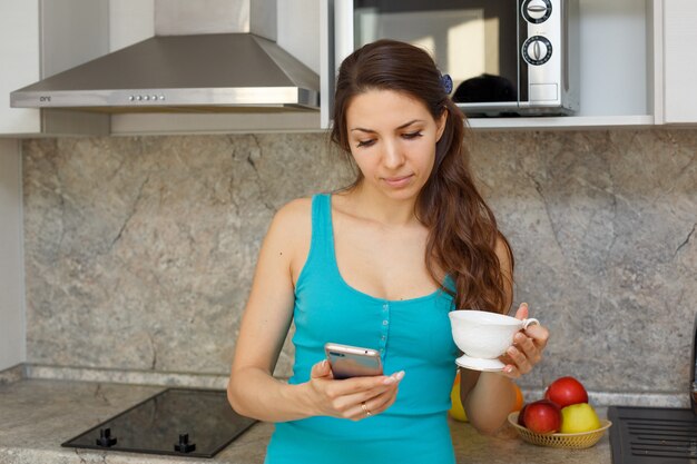 Una linda mujer con una camiseta verde y cabello oscuro está parada en la cocina con una taza y un teléfono en la mano.
