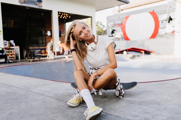 Linda mujer caucásica posando con placer en el parque de patinaje. Atractiva joven rubia sentada en patineta y sonriendo.