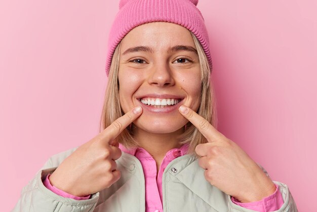 Linda mujer caucásica positiva señala con el dedo índice una sonrisa con dientes que muestra sus dientes perfectos, usa sombrero y chaqueta, está de buen humor aislado sobre un fondo rosado Concepto de personas y felicidad